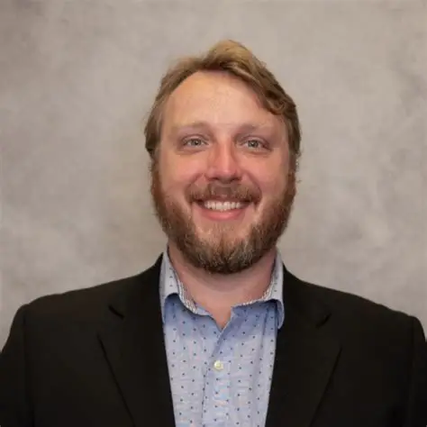 Smiling man with a beard wearing a black blazer and a blue patterned shirt.