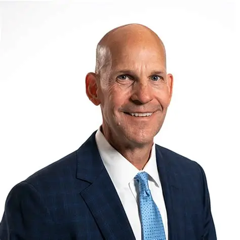 Smiling bald man in a navy blue suit and patterned tie against white background.