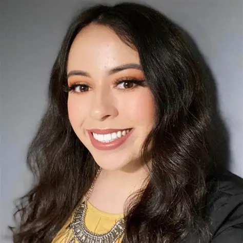 Smiling woman with long dark wavy hair, wearing a yellow top and silver necklace.