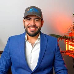 A smiling man with a beard wears a blue blazer and a grey mountain-logo baseball cap.