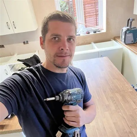 Man holding a power drill in a kitchen renovation with white cabinets and wooden countertops.