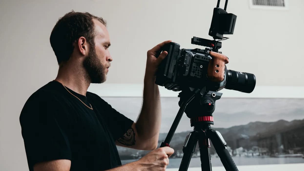 Bearded man in a black shirt operates a professional cinema camera mounted on a tripod.