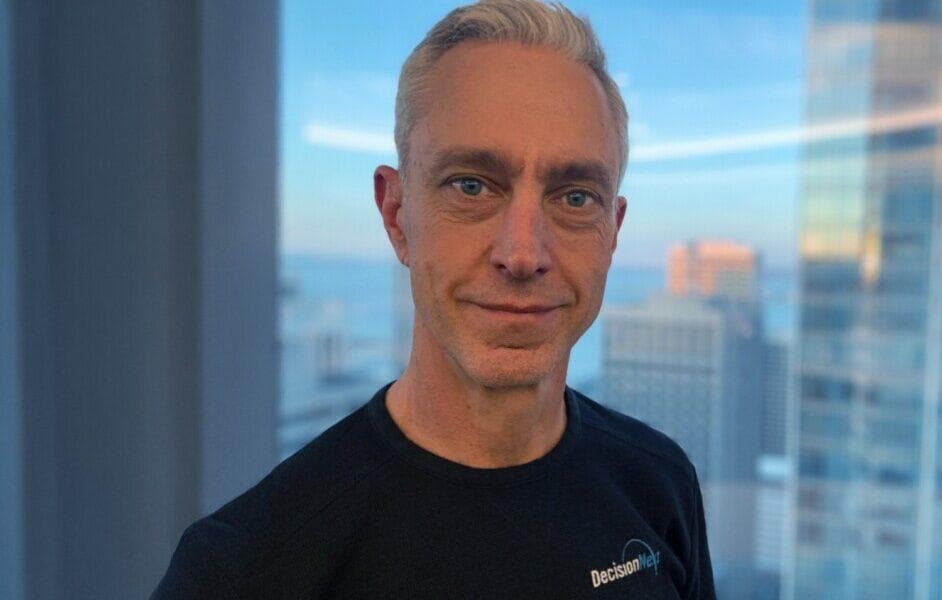 Smiling man with gray hair in a black shirt, with a blurred cityscape background.