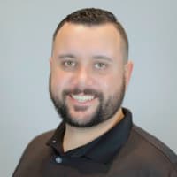 Smiling man with a beard wearing a black polo shirt against a plain gray background.