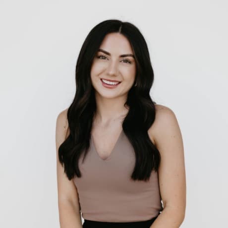 Smiling woman with long dark hair wearing a mauve sleeveless top against a white background.